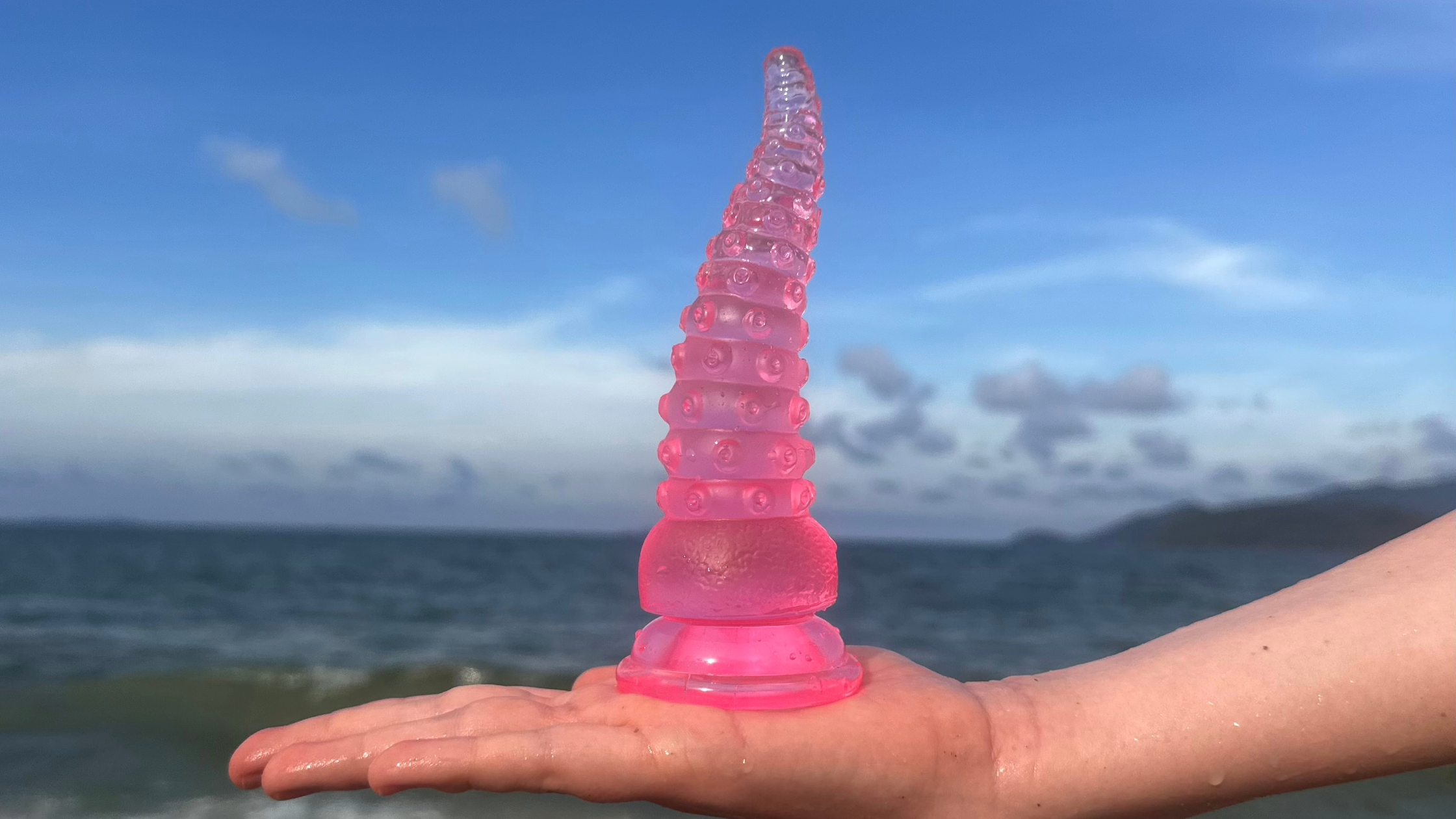 Pink Fantasy Dildo on hand with beach in the background Numen Wellness